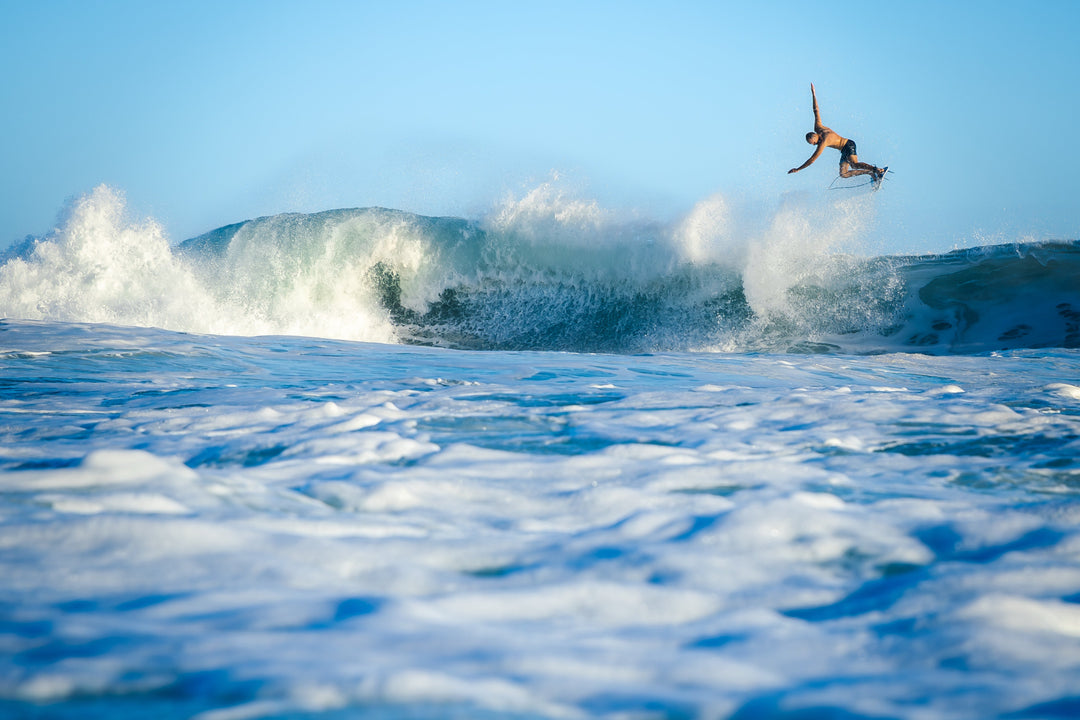 A shirtless male surfer launches high into the air off the lip of a large crashing wave, photographed from water level against a clear blue sky.