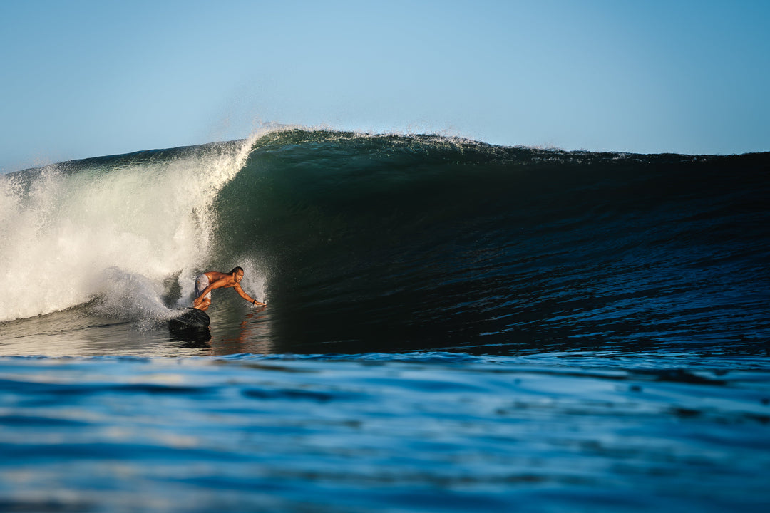 A shirtless man carves along the face of a massive barreling ocean wave, photographed from water level with spray cascading off the lip above him.