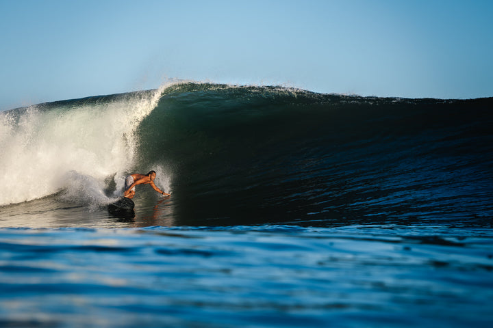 A shirtless man carves along the face of a massive barreling ocean wave, photographed from water level with spray cascading off the lip above him.