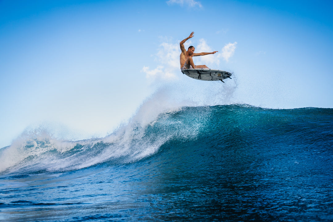 Male surfer Flynn Novak launches a high aerial off the lip of a barreling blue ocean wave under a clear sky, wearing Sandy Brushstroke board shorts.