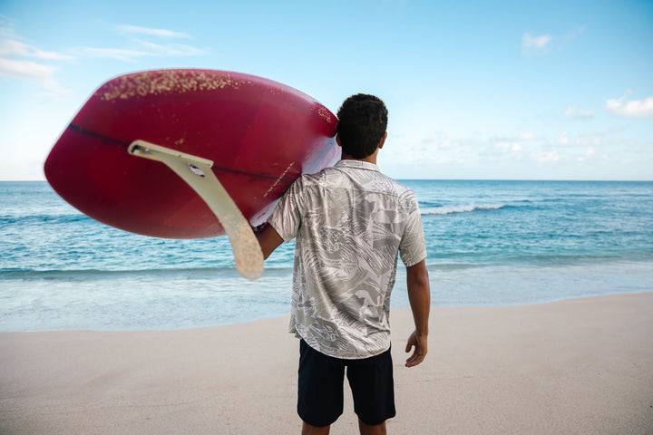 Man viewed from behind carrying a red surfboard on his shoulder while standing on a sandy shore, wearing a light patterned shirt and dark shorts, gazing at the turquoise ocean.