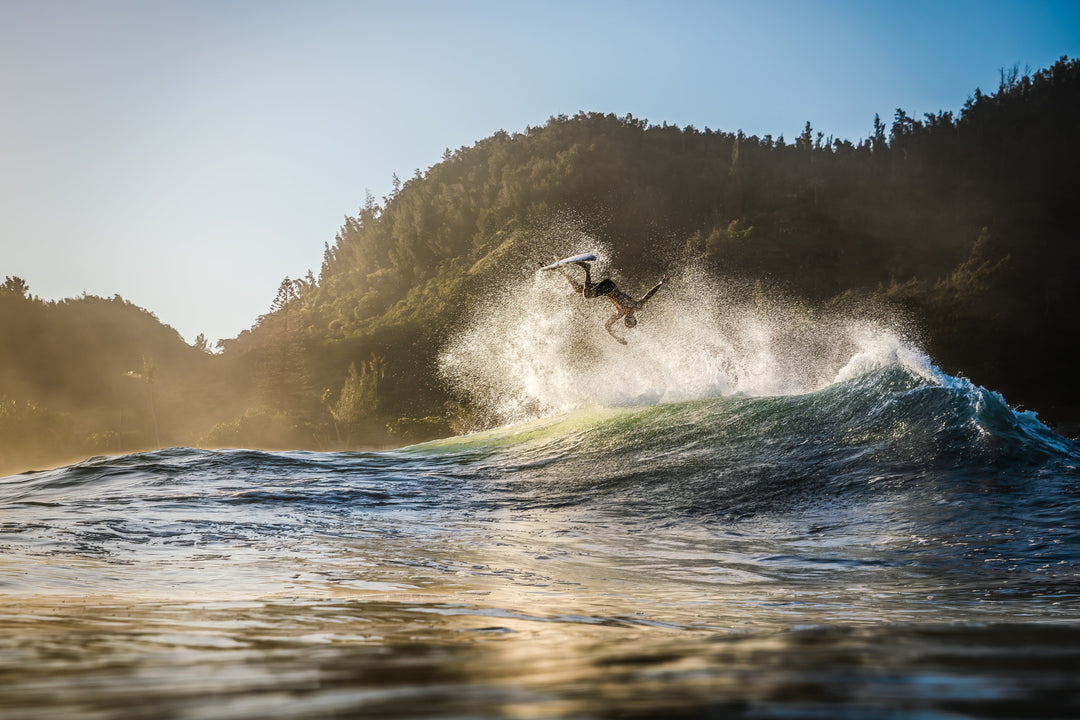 A surfer executes a dramatic aerial trick off a glassy green wave, surrounded by white spray with a tree-covered hillside and golden haze in the background.