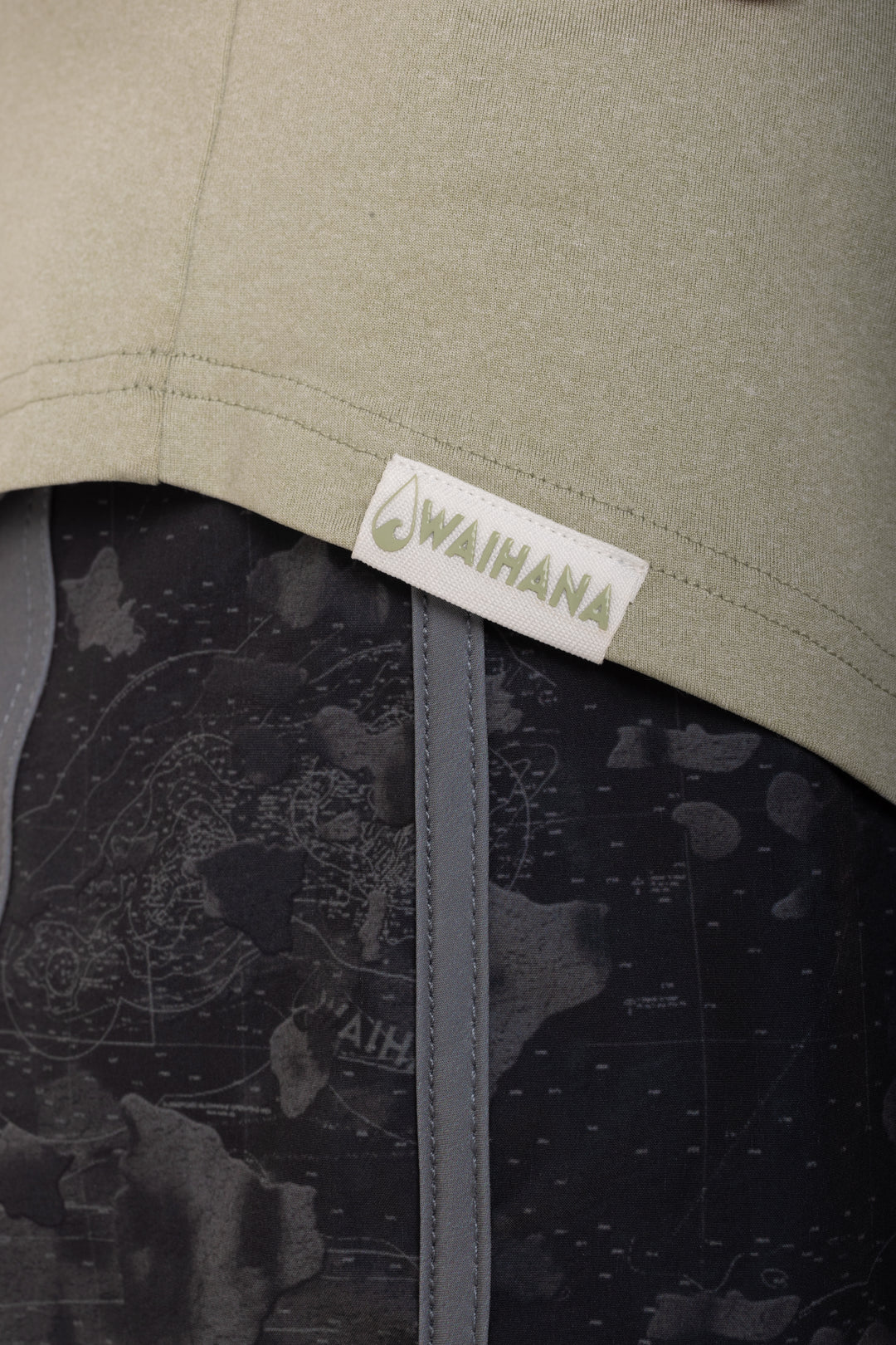 Close-up of a woven Waihana brand label stitched into the hem of the Mariner Green Hooded Long Sleeve sun shirt, with dark map-print shorts visible beneath.