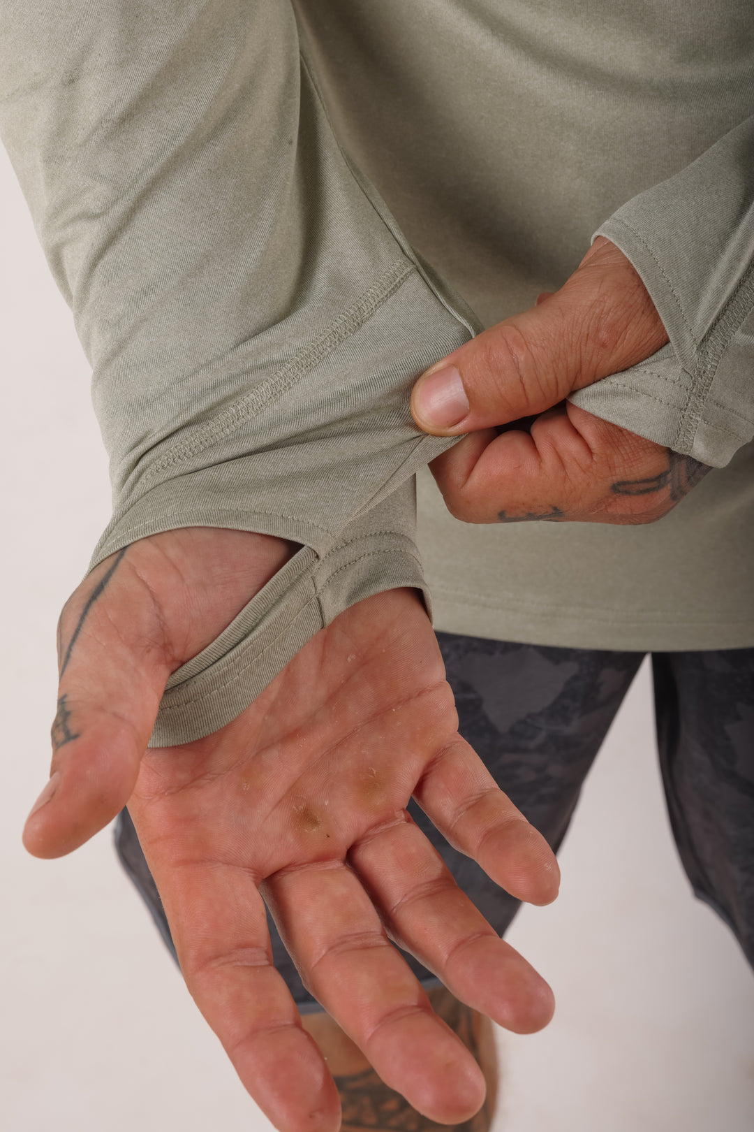 Close-up of tattooed hands pulling back the sleeve of a sage green Waihana Mariner hooded long-sleeve sun shirt to show the thumb hole cuff feature.
