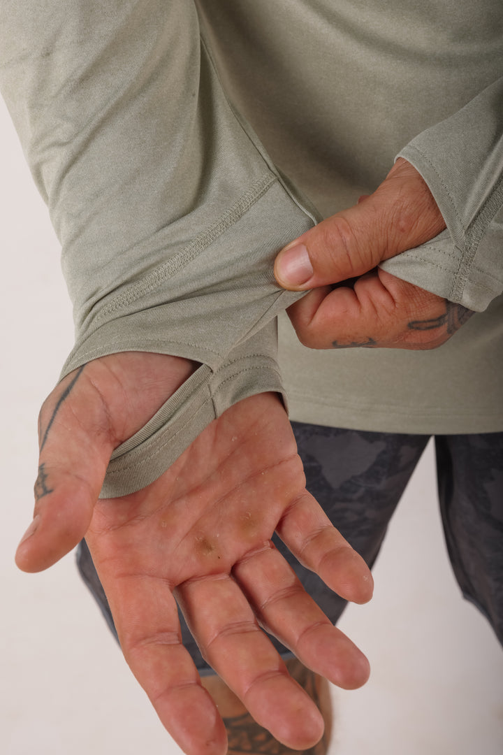 Close-up of tattooed hands pulling back the sleeve of a sage green Waihana Mariner hooded long-sleeve sun shirt to show the thumb hole cuff feature.