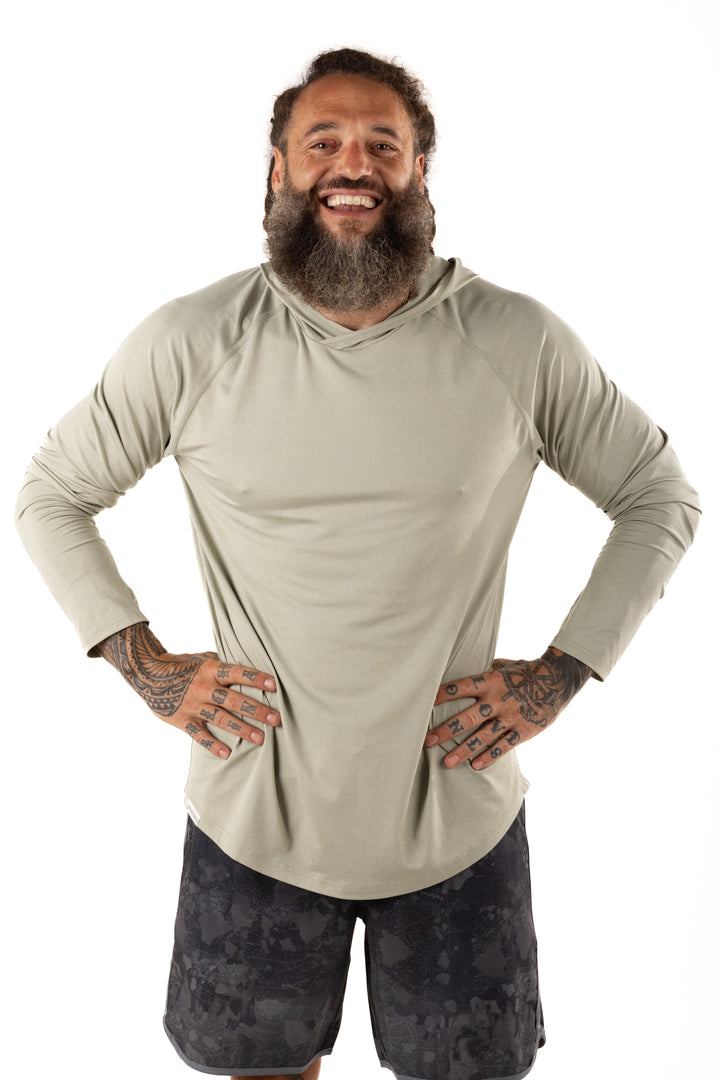Smiling tattooed man wearing the Waihana Mariner Green Hooded Long Sleeve sun shirt with dark patterned board shorts, hands on hips against a white studio background.