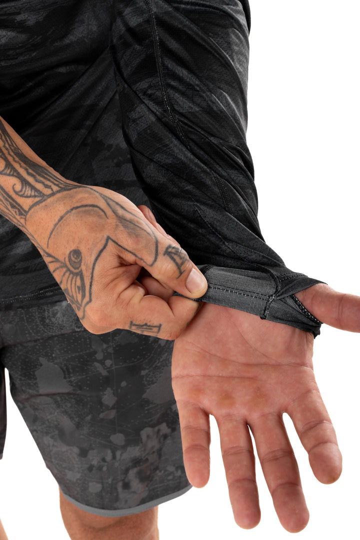 Tattooed man stretching the thumb loop wrist cuff of the Waihana Tropicam Black long-sleeve sun shirt, highlighting the secure fit detail against a white background.